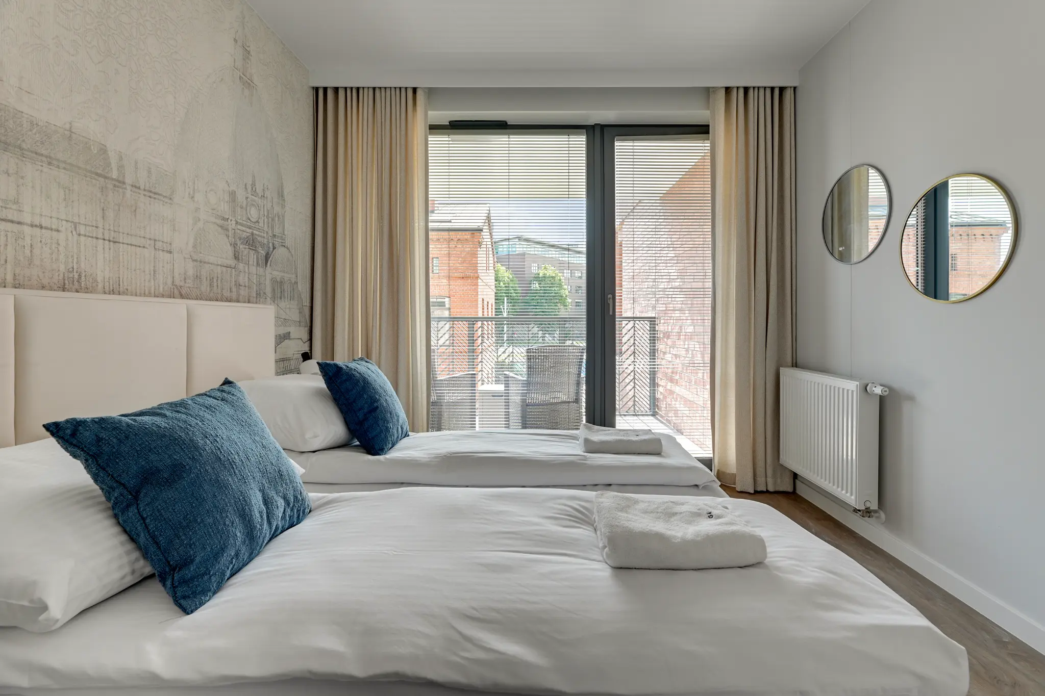 A bedroom in a Scala apartment with two beds, architecture-patterned wallpaper and glazing onto the balcony