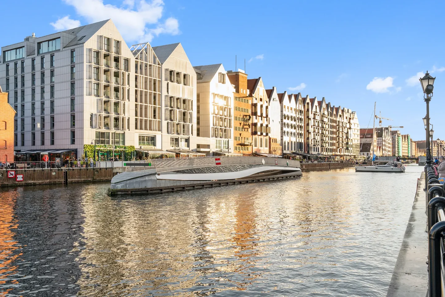 A view across the water towards a row of tall buildings with pitched roofs and a low footbridge.