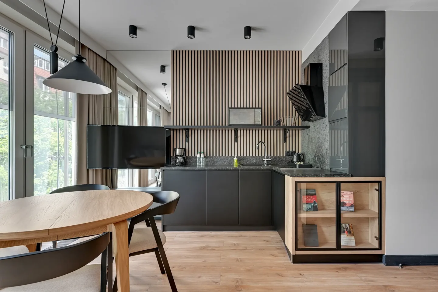 Kitchenette with graphite cabinets, wooden slats and a round table beside large windows.