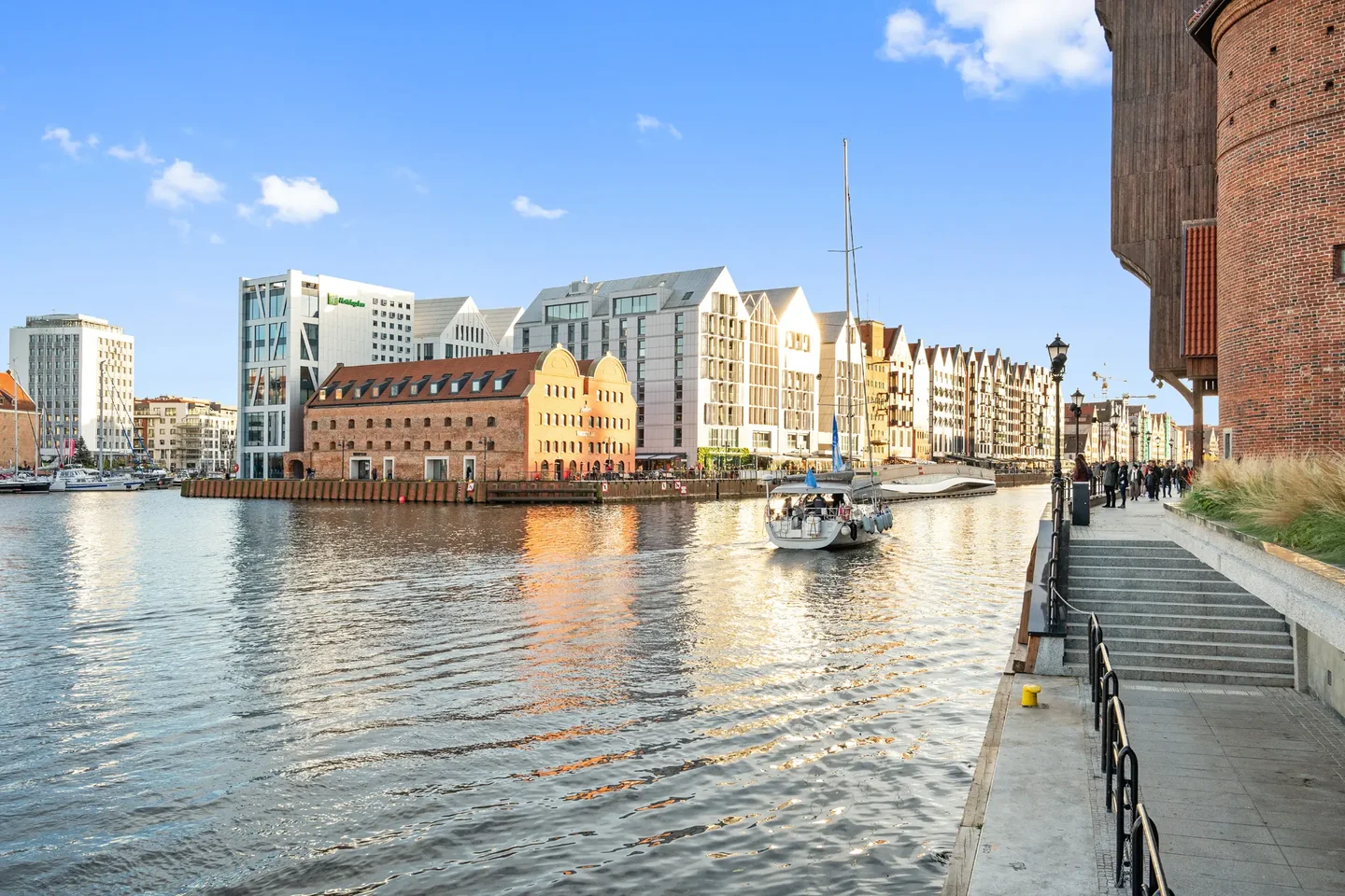A sailing boat on the water between built-up waterfronts, beside a promenade with steps and a brick building.